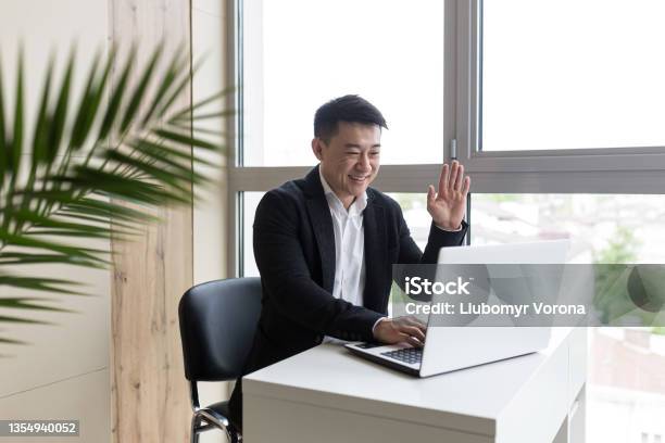 Successful Asian Businessman In A Black Business Suit Works On A Laptop In A Stylish Office Explains A New Business Strategy Via Video Link Stock Photo - Download Image Now