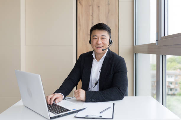 male businessman conducts online training using a headset and laptop, sits in a stylish office by the window and looks at the camera friendly - friendly match imagens e fotografias de stock