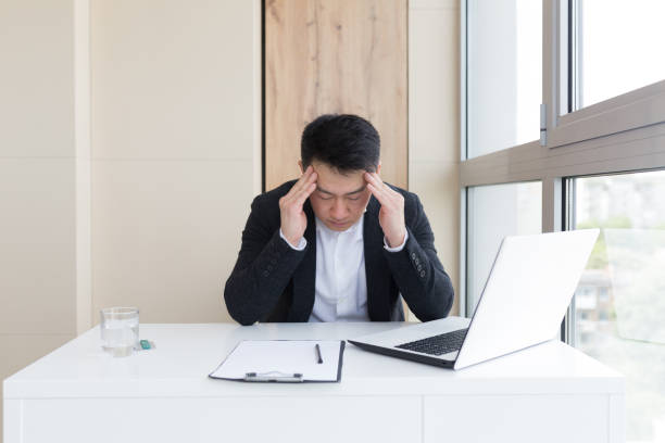 istock young asian office worker suffering from headache at work in the office drinks medicine pill with water. Sick man in a suit at the computer indoors with severe pain ache uses the drug in workplace 1354940136