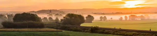 Panoramic view over misty morning valley to distant city and cathedral silhouetted by the golden light of sunrise.