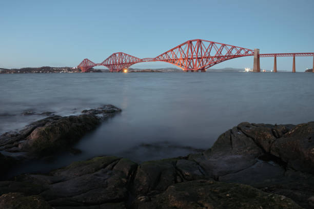 vue sur le pont ferroviaire du forth en soirée et la côte rocheuse au premier plan - rivière firth of forth photos et images de collection