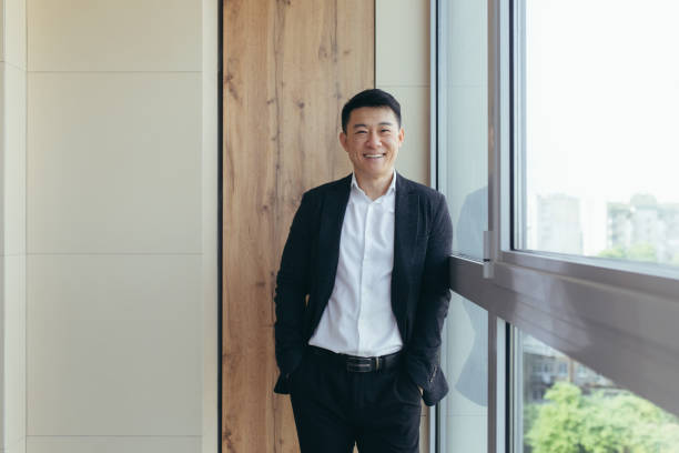 Freelancer Asian businessman in business suit looking at camera near the window of a modern office center Freelancer Asian businessman in business suit looking at camera near the window of a modern office center men standing business formalwear stock pictures, royalty-free photos & images