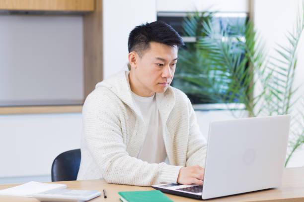 istock Young asian man working at home on a laptop computer sitting at a table in the kitchen 1354944041
