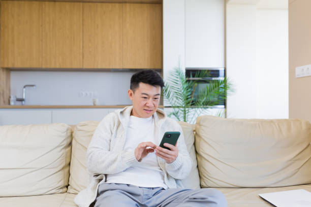 istock Asian man sitting at home on the couch, using a smartphone, serious typing a message on the phone 1354944347