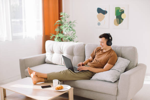 istock Relaxed young guy in wireless headphones lying on sofa in minimalistic living room and using laptop while working online 1354947704