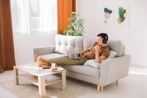 istock Relaxed young guy in wireless headphones lying on sofa in minimalistic living room and using laptop while working online 1354947710
