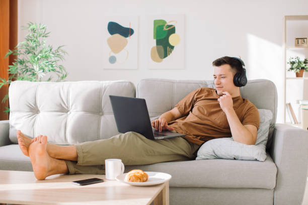 istock Relaxed young guy in wireless headphones lying on sofa in minimalistic living room and using laptop while working online 1354947711