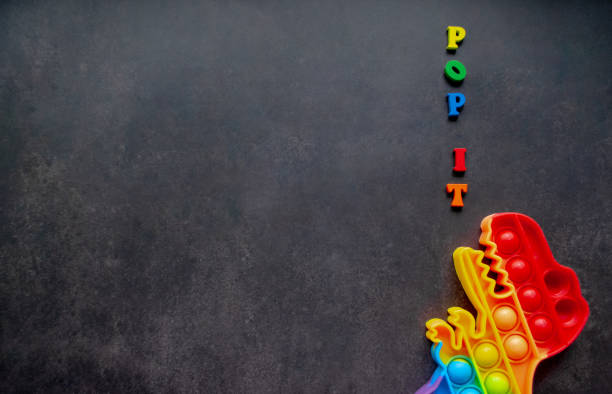 istock Toy pop it dinosaur rainbow colors on a black background with multicolored letters and lettering - Pop it. Copy space 1354948178