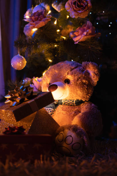 istock Teddy bear holding a gift box with glowing light under a Christmas tree in the dark. 1354957480