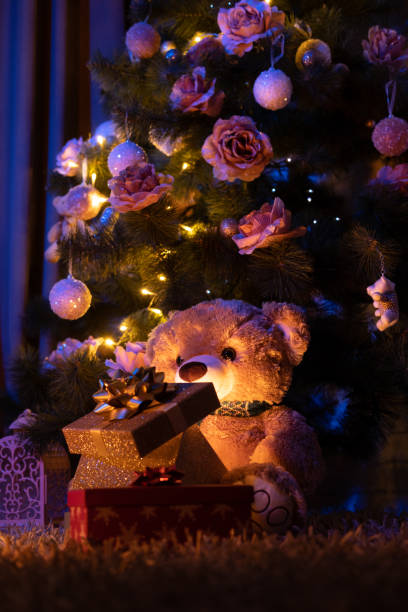 istock Teddy bear looking in an opened gift box with light shining out, sitting under a Christmas tree. 1354957587