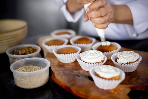 Close-up on a baker decorating some cupcakes at a pastry shop using whipped cream