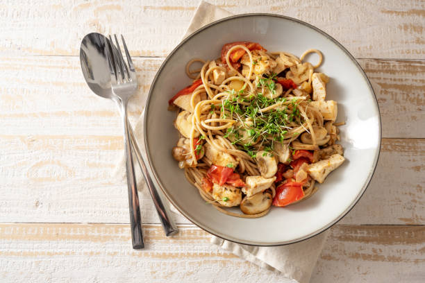 istock Whole grain spaghetti with tomato, mushroom and chicken strips, garnished with cress, gray plate and cutlery on a light rustic wooden table, top view from above 1354963628