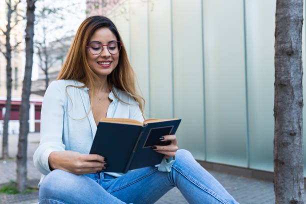 istock Positive optimistic brunette woman with glasses smiling while reading book outdoors. Student concept 1354975079