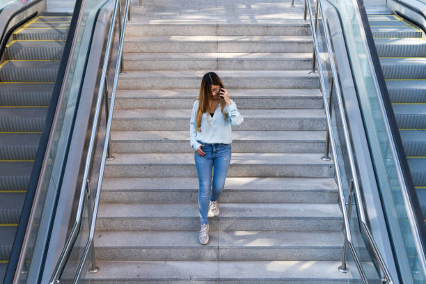 istock Young businesswoman with glasses using smartphone while smiling, going down on metro, airport stairs. Business concept 1354975637