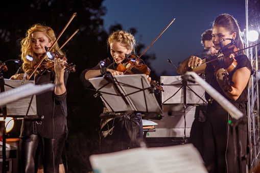 Charming women violinists looking at musical notes and playing melody on violin while performing outdoor concert under night sky