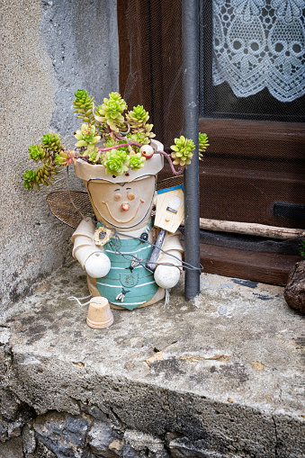 cute flowerpot in the shape of a doll dressed as a farmer built with other flowerpots, with sempervivum plants on its head, placed in the window sill of a rural house, vertical