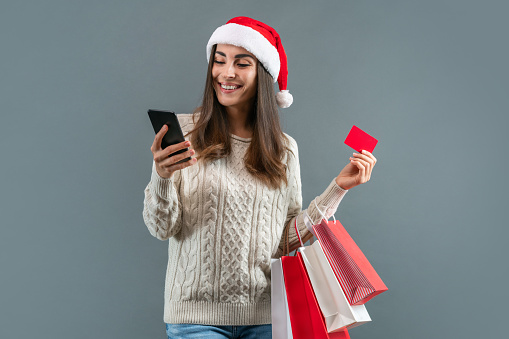 Christmas and new year sales, holiday shopping concept. Attractive smiling girl in Santa hat with a bunch of shoppers and credit card in hands making online order via her mobile phone.