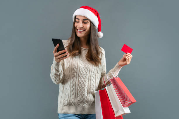 attractive smiling girl in santa hat with a bunch of shoppers and credit card in hands making online order via her mobile phone - shopping e commerce internet credit card imagens e fotografias de stock