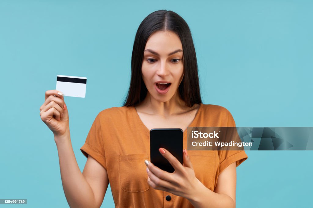 Portrait of an excited young brunette girl showing plastic credi Portrait of an excited young brunette girl showing plastic credit card while browsing mobile phone, isolated over pastel blue background. Adult Stock Photo