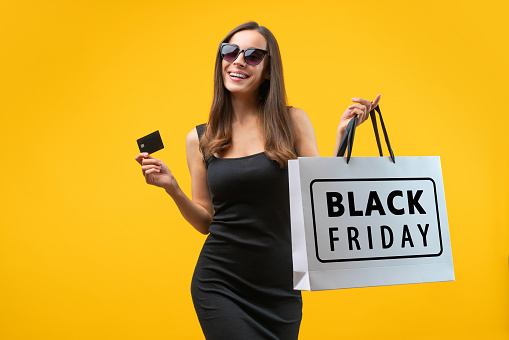 Studio portrait of attractive confident young woman in trendy black dress and glasses, posing over bright colored yellow background with credit card and Black Friday shopping bag in hands.