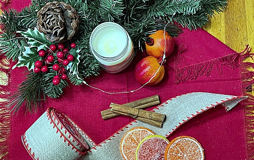 Holiday flat lay grouping of pine boughs around scented candle near cinnamon sticks and candied citrus on burlap ribbon.