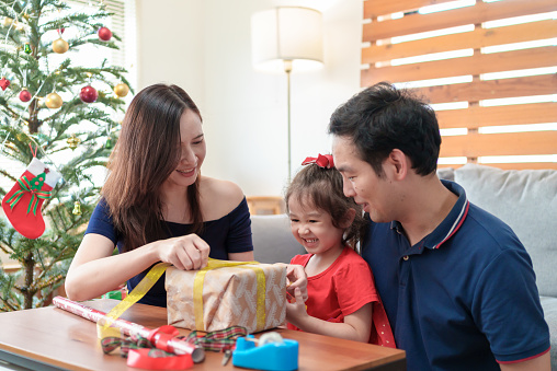 Young Asian parents and adorable daughter making gift box together at home, preparing for Christmas. Happy Family decorating handmade Christmas presents with ribbon together