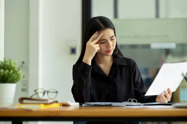 Thoughtful and stressed-out businesswoman examines financial report paperwork at her office desk.