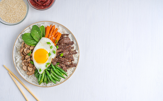 Directly above view of Asian Bibimbap dish made of boiled rice topped with beef slices, vegetables and fried egg with yolk served on plate with chopsticks on white wooden background with copy space