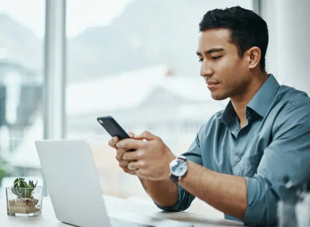 Photo of Shot of a young businessman using a smartphone and laptop in a modern office