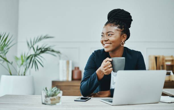 photo d’une jeune femme d’affaires utilisant un ordinateur portable et prenant un café dans un bureau moderne - technology contemplation clothing formalwear photos et images de collection