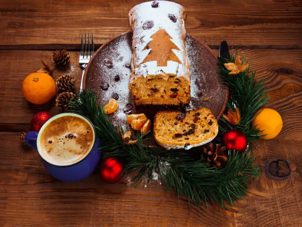 Christmas fruitcake with powdered sugar fir tree decoration and coffee cup tangerines cranberries festive holiday wooden table flat lay. Stollen cake loaf Wholegrain flour dough baking recipe top view