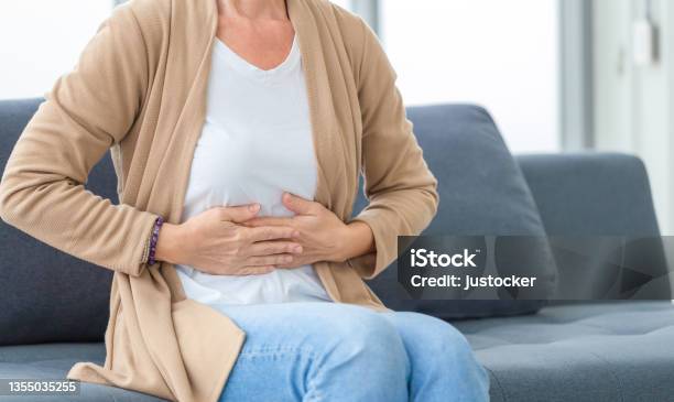 Unhappy Woman Stomach Ache Mature Woman With Stomach Pain Feeling Unwell Sitting In Living Room Stock Photo - Download Image Now