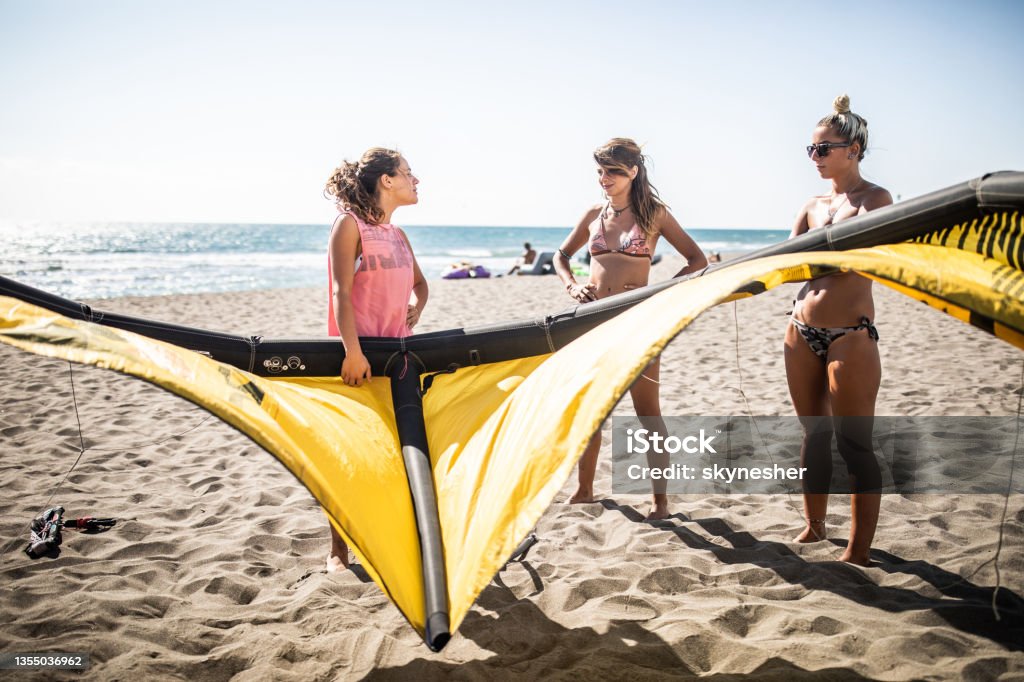 Group of young women talking while preparing for kitesurfing on the beach. Three female friends preparing their equipment for kiteboarding in summer day on the beach. Kiteboarding Stock Photo