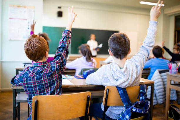 Schoolchildren at classroom with raised hands answering teacher's question. stock photo