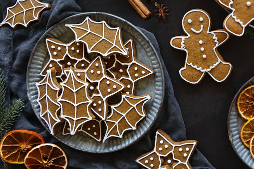Stock photo showing close-up, elevated view of cooling rack of homemade gingerbread men and plate of holly leaf and star biscuits on black background with star anise, spruce needles, dried orange citrus slices, red berries and cinnamon sticks. Home baking concept.