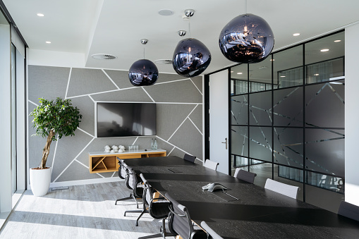Black conference table, surrounding chairs, sculptural pendant lights, potted plant, sideboard, and LCD television in sunny meeting room.