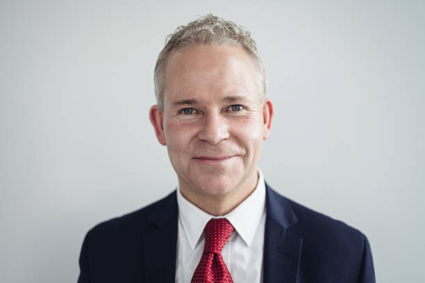 Indoor portrait of cheerful businessman in corporate attire Head and shoulders view of executive in mid 50s wearing white shirt, red tie, and blue suit jacket smiling at camera against gray background. headshot stock pictures, royalty-free photos & images