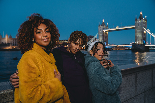 Portrait of three young adult hip friends in London near Tower Bridge. Night time. They are wearing baggy hip and colorful clothes.