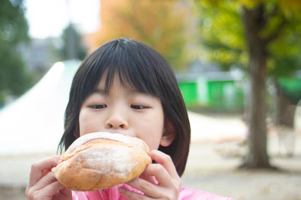Japanese girl is eating a large bread outdoors. She has it by both hands and looking it.
