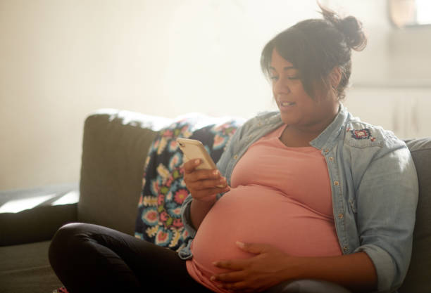 photo recadrée d’une jolie jeune femme enceinte en ensençant un texto alors qu’elle était assise dans son salon à la maison - human pregnancy photos et images de collection