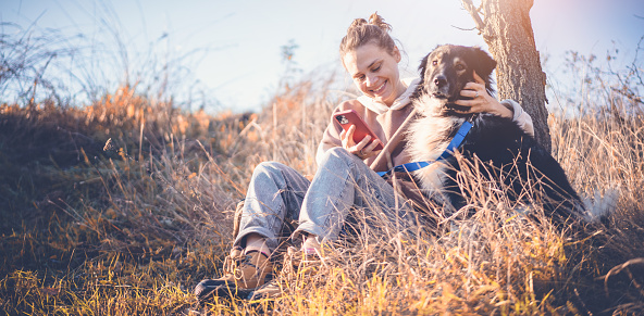 Beautiful happy young woman sitting on the grass hugging a dog with a smartphone in her hands. Love and affection for pets, friendship companion