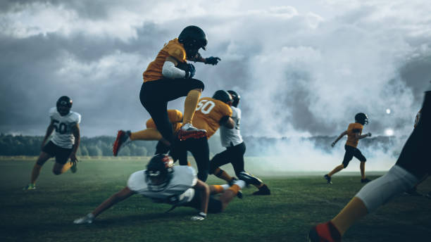 campo de futebol americano duas equipes competem: jogador bem sucedido saltando sobre a defesa correndo para marcar pontos de touchdown. atletas profissionais competem pela bola, enfrentam, lutam pela vitória no campeonato - football player group of people running american football - fotografias e filmes do acervo