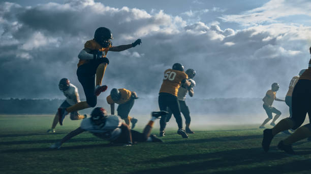 campo de futebol americano duas equipes competem: jogador bem sucedido saltando sobre a defesa correndo para marcar pontos de touchdown. atletas profissionais competem pela bola, enfrentam, lutam pela vitória no campeonato - football player group of people running american football - fotografias e filmes do acervo