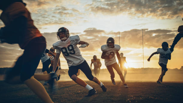 american football field two teams compete: players pass and run attacking to score touchdown points. professional athletes fight for the ball, tackle. golden hour sunset shot - playback imagens e fotografias de stock