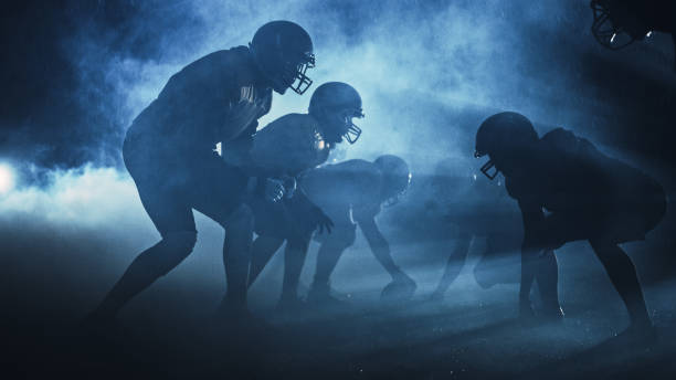 campo di football americano due squadre competono: i giocatori passano, corrono, attaccano per segnare punti touchdown. notte piovosa con atleti che combattono per la palla in fumo drammatico. - scuro immagine foto e immagini stock