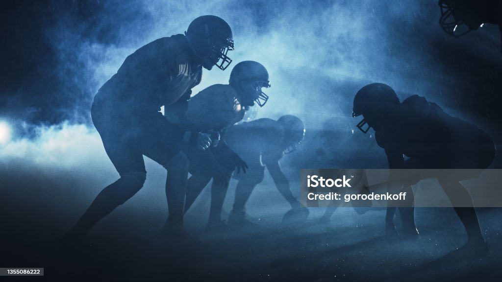 American Football Field Two Teams Compete: Players Pass, Run, Attack to Score Touchdown Points. Rainy Night with Athletes Fight for the Ball in Dramatic Smoke. American Football - Sport Stock Photo