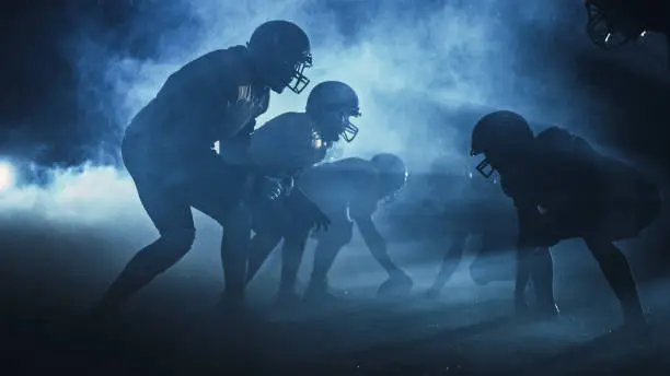 Photo of American Football Field Two Teams Compete: Players Pass, Run, Attack to Score Touchdown Points. Rainy Night with Athletes Fight for the Ball in Dramatic Smoke.