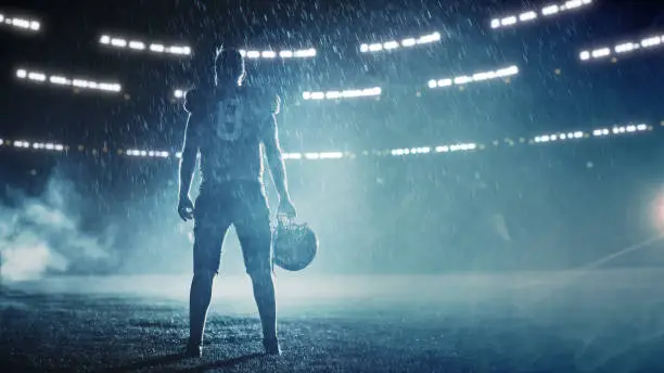 Photo of American Football Staduim: Lonely Athlete Warrior Standing on a Field Holds his Helmet and Ready to Play. Player Preparing to Run, Attack and Score Touchdown. Rainy Night with Dramatic Fog, Blue Light