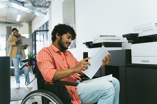 Mixed race worker in modern office coworking place. Disabled indian businessman making photo copies of documents wheelchair.