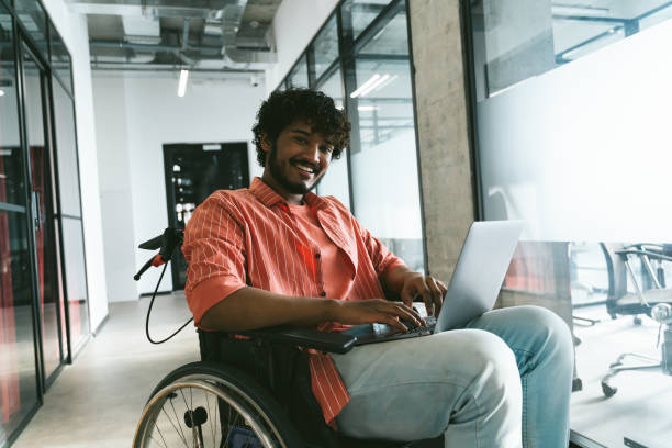Disabled mixed race indian office worker in modern office corridor using laptop. Businessman in wheelchair
